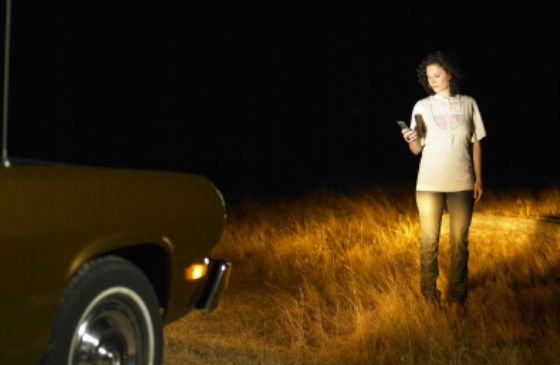 Woman on the side of the road, using her cell phone beside a broken-down car.