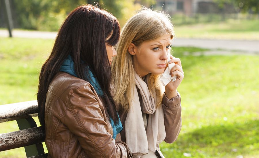 Two women sit on a park bench, as one comforts the other.