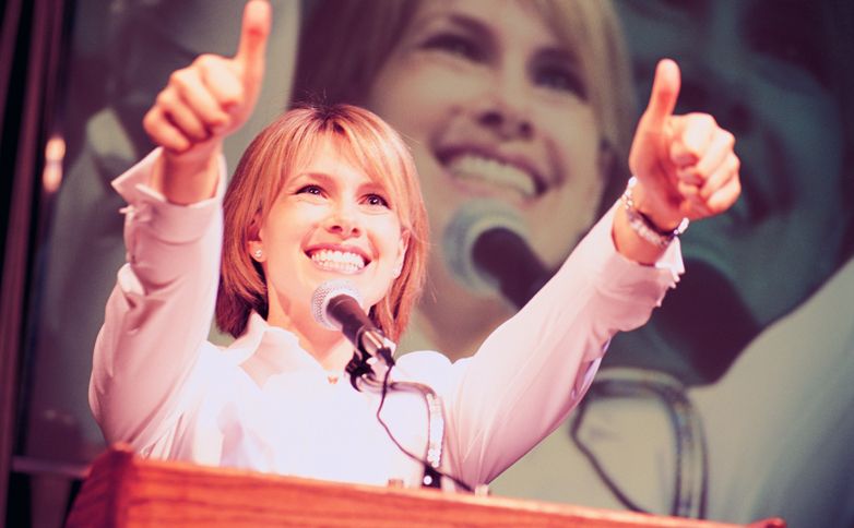 A proud, confident woman speaks before a large, attentive audience.