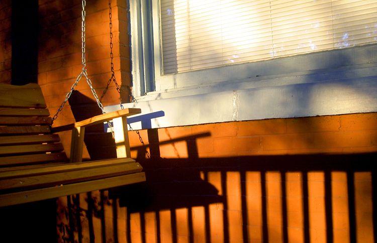 A wooden swing sways in the dappled sunlight of a porch.