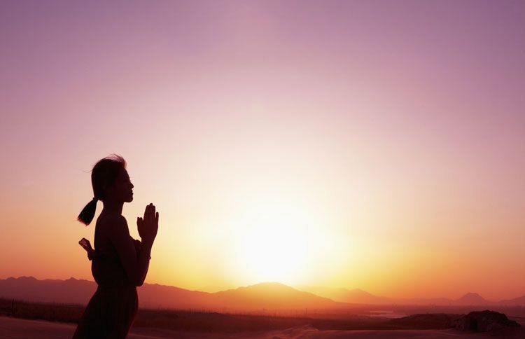 A woman stands in prayer as the sun sets in the background