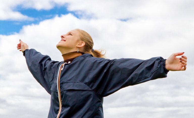 A smiling woman stands, arms spread, gazing up at the sky