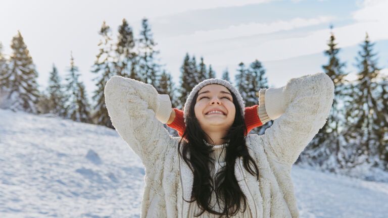 Woman in a coat and hat outside in the snow smiles with eyes closed and says a winter prayer