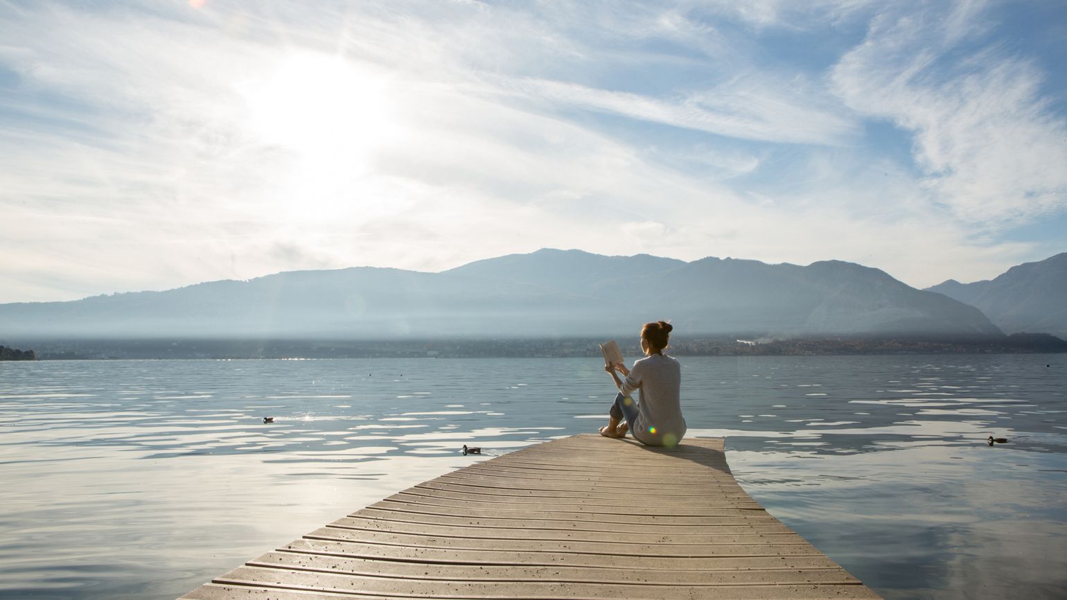 Young woman sits on a jetty above the lake, she is reading a book. Beautiful Autumn day in Italy