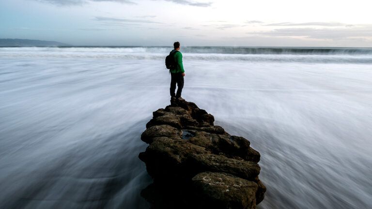 A man stands upon a rock while water rushes by on both sides.