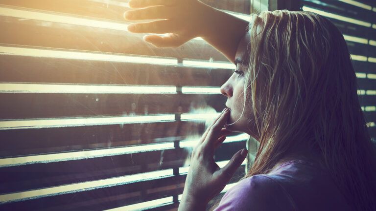 A grieving woman looks out her window through the blinds