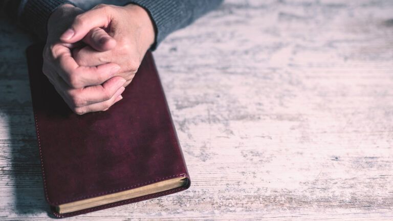 A man prays, his clasped hands resting on a Bible