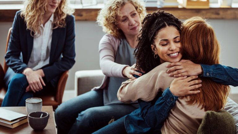 Caring female counselor hugs a female patient during a group therapy session.